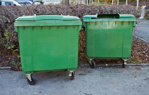 Two large green wheeled waste bins positioned side by side on a paved surface in an outdoor setting, with a background of leafless bushes and a pathway. The larger bin on the left and the smaller bin on the right are both made of sturdy plastic material with smooth, slightly glossy finishes. Each bin has a hinged lid on top, with the larger bin's lid fitting snugly, while the smaller bin's lid appears slightly ajar. The bins are placed on the ground without any visible contents, indicating they are ready for rubbish collection. The environment suggests a residential or commercial area, consistent with local rubbish clearance services in Ruislip, where Garden Clearance Ruislip provides waste removal. The scene reflects a typical garden or property waste disposal setup, emphasizing cleanup or clearance operations within the local postcode area.