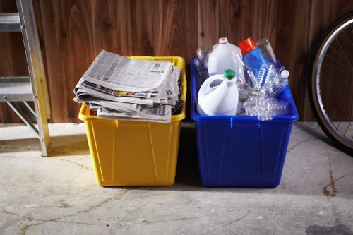 Inside a garage or storage area with a wooden wall and concrete floor, two large plastic recycling bins are positioned side by side. The yellow bin on the left contains a stack of flattened newspapers and magazines, with some overlapping and protruding slightly beyond the top edge. The blue bin on the right holds various household plastics, including clear plastic bottles, a white detergent bottle with a green cap, and some empty, crushed plastic containers. The bins are situated against the wall, with a portion of a metal shelving unit visible to the left and a bicycle wheel partially seen on the right side of the image. The scene suggests an organized approach to waste sorting, typical of household or small-scale waste management, and aligns with the services provided by Garden Clearance Ruislip in managing rubbish and recycling needs in the local area near Ruislip, Hillingdon ward.