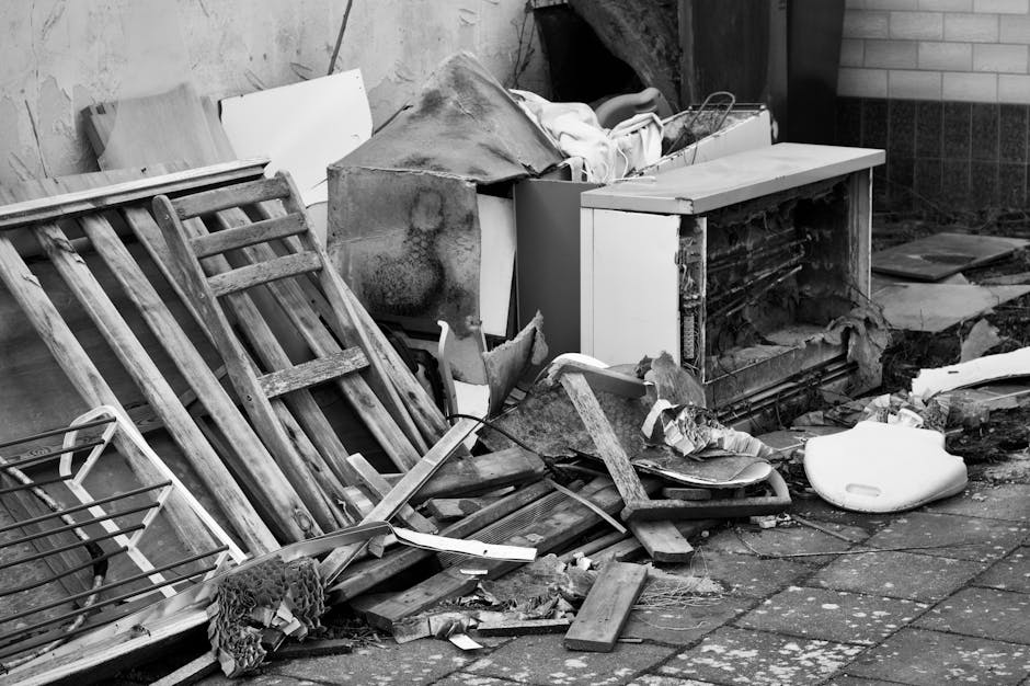 A photograph depicting a large pile of mixed household and garden waste materials stacked in an outdoor area, likely a driveway or cleared space adjacent to a building. Prominent in the foreground are