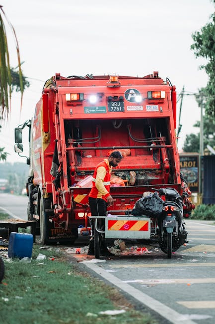 A street scene showing a white commercial garbage truck with an open rear loading compartment, positioned alongside a sidewalk in an urban environment. The truck's rear is equipped with mechanical arm