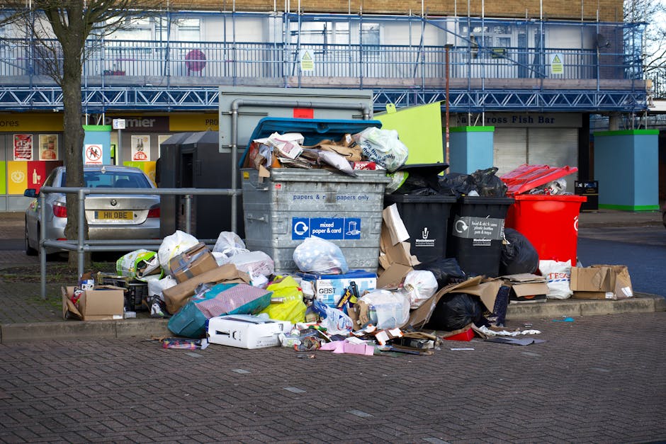 A collection of overflowing rubbish bins and loose waste bags are piled on the pavement in an urban area, with various cardboard boxes, plastic bags, and discarded packaging scattered around the base.
